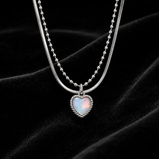 A double-strand silver necklace featuring a textured snake chain and a ball chain with a heart-shaped iridescent opal pendant, displayed on a black velvet background.